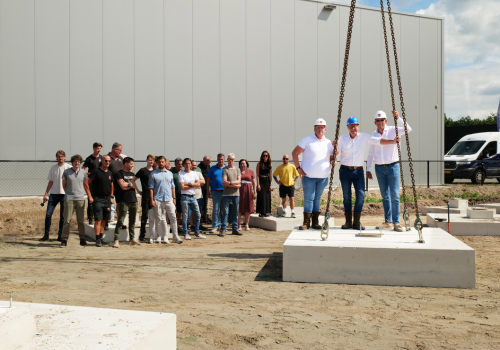 Groep mensen bij bouwplaats, drie personen met helm staan op betonnen funderingselement tijdens symbolische start bouw